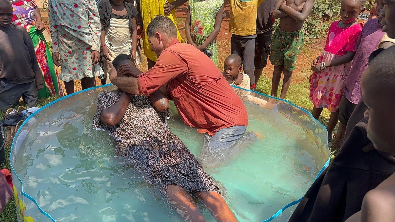 A group of people gathered around a small, makeshift pool where a person in a leopard-print dress is in the water, receiving a religious baptism from a man in an orange shirt. Several onlookers, including children and adults, watch the scene outdoors on a sunny day.
