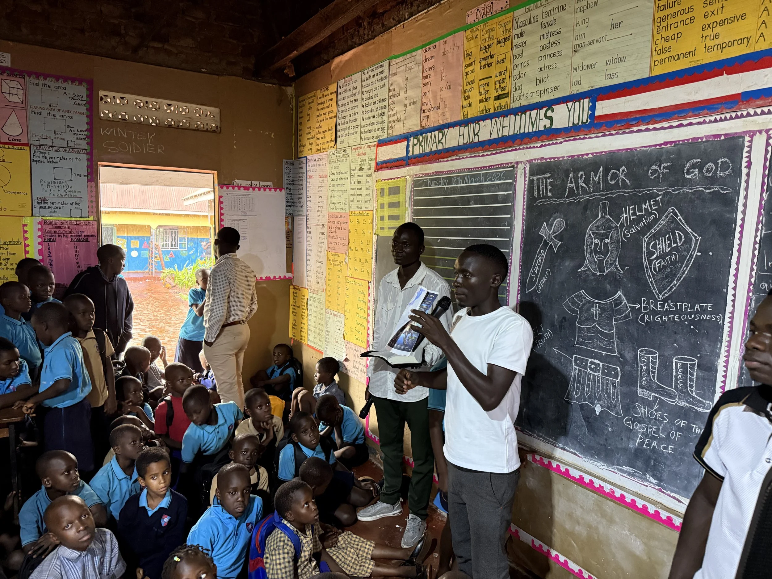 Classroom with students in blue uniforms sitting on the floor as teachers or volunteers speak at the front. Chalkboard with drawings of armor and signs, colorful posters and educational charts on the walls, some students looking attentively, sunlight coming through open doorway.