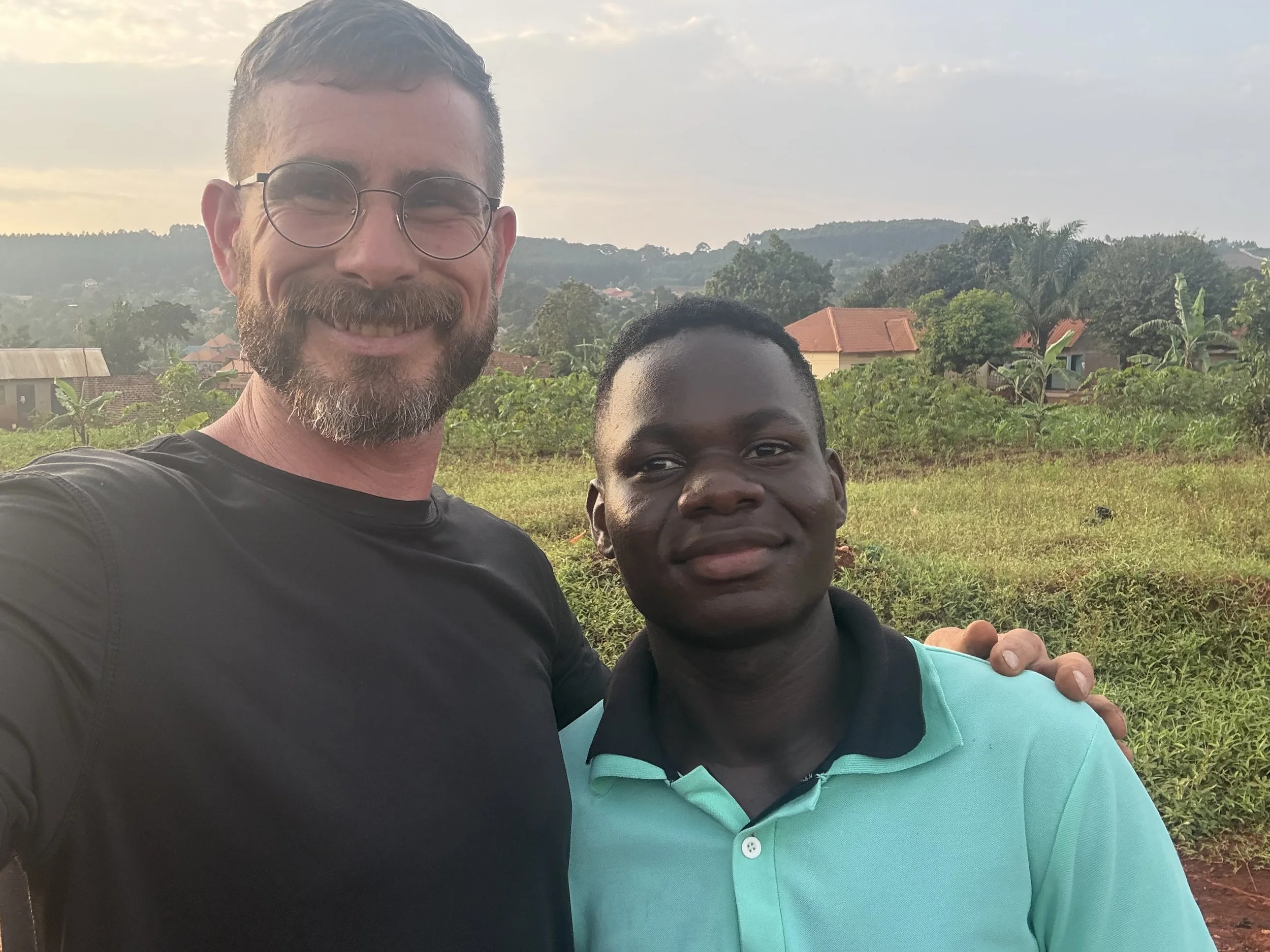 A man with glasses and a beard smiling next to a boy in a light blue shirt, outdoors with houses and greenery in the background.