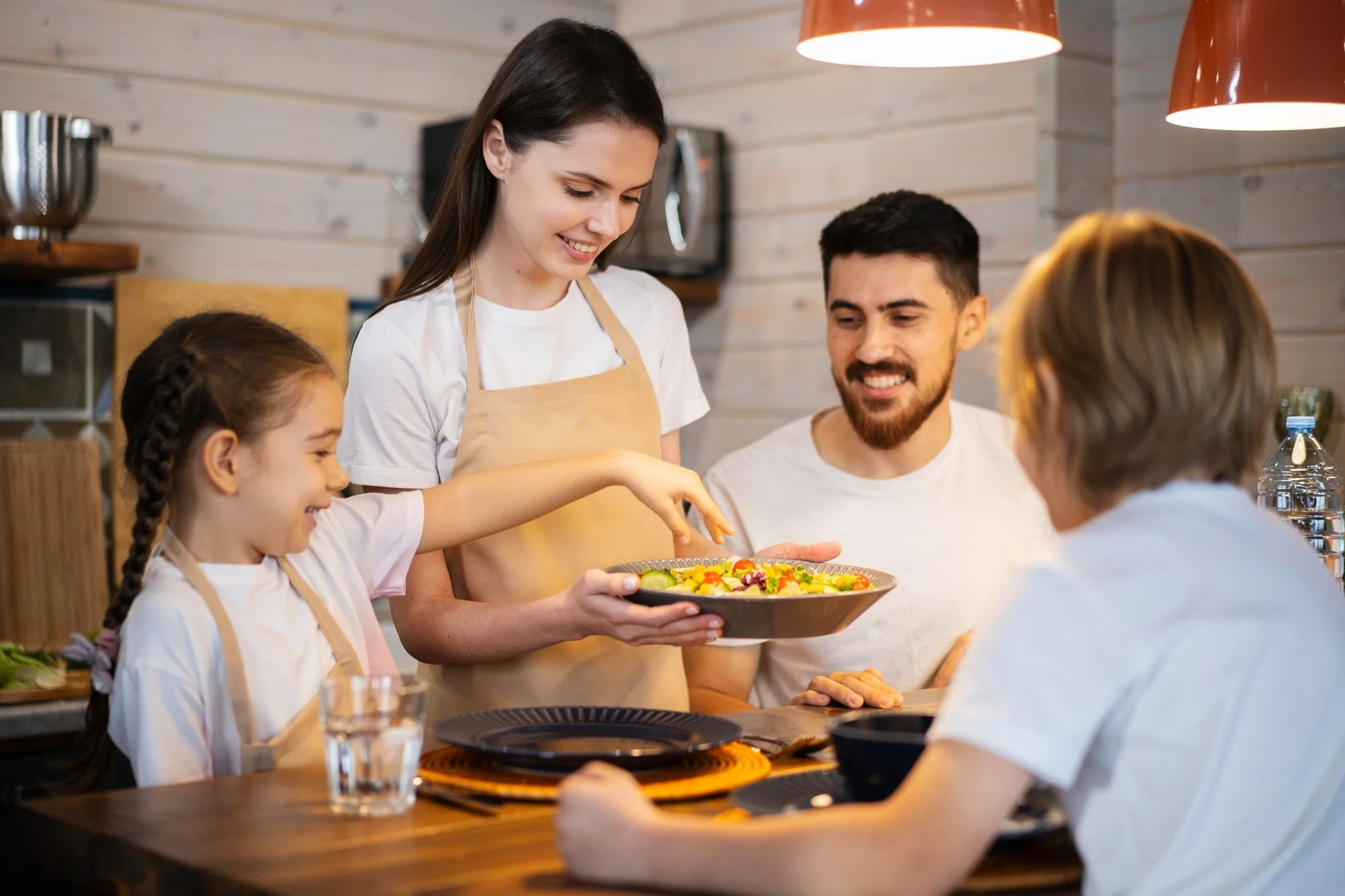 Family enjoying a meal together in a cozy kitchen, with a woman serving a colorful salad to a young girl, while a man and another child sit at the table.