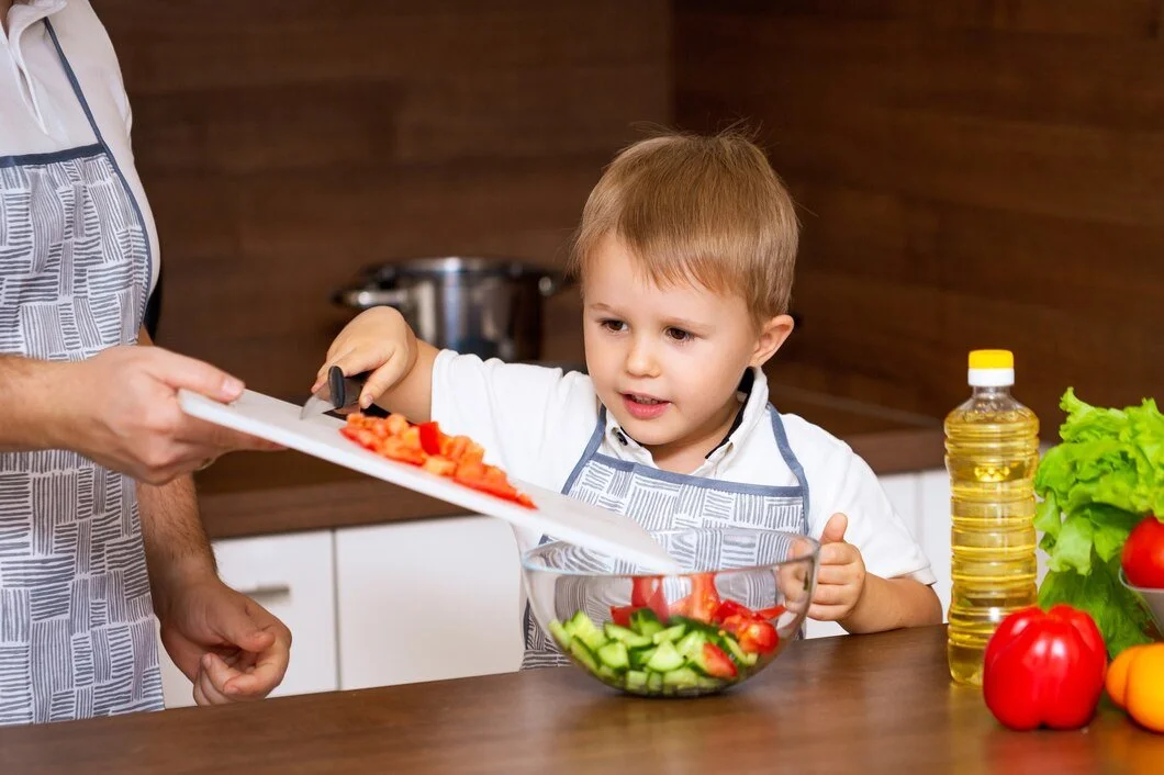 A young boy is watching an adult cut tomatoes and place them into a glass bowl filled with cucumbers and tomatoes. The table has fresh vegetables, a bottle of cooking oil, and lettuce.