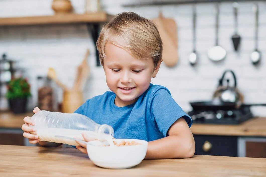 A young boy in a blue shirt pouring milk from a plastic bottle into a bowl of cereal in a modern kitchen.