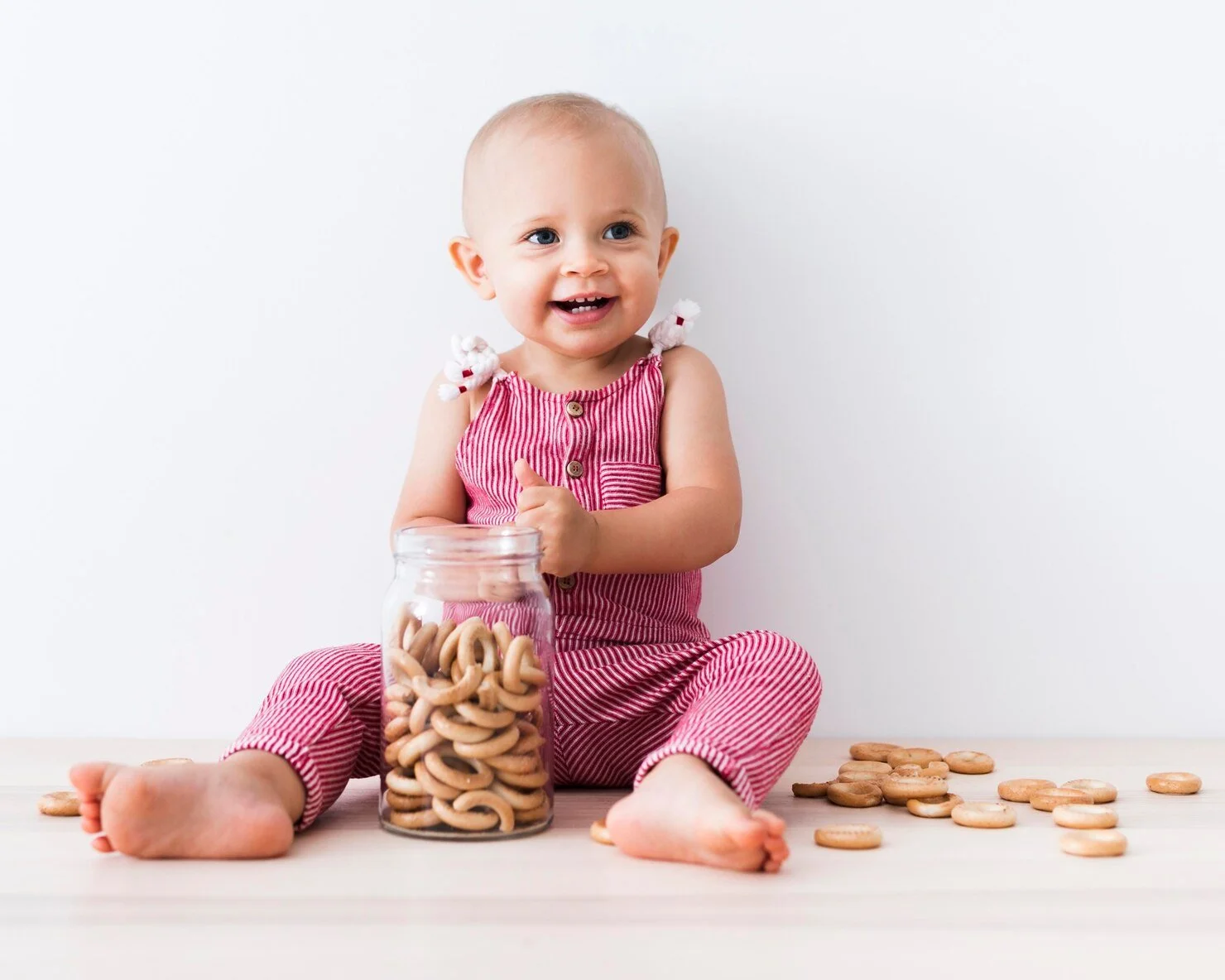 A smiling baby girl dressed in a pink and white striped outfit, sitting on a wooden floor with a jar of cookies in front of her and some cookies scattered on the floor.