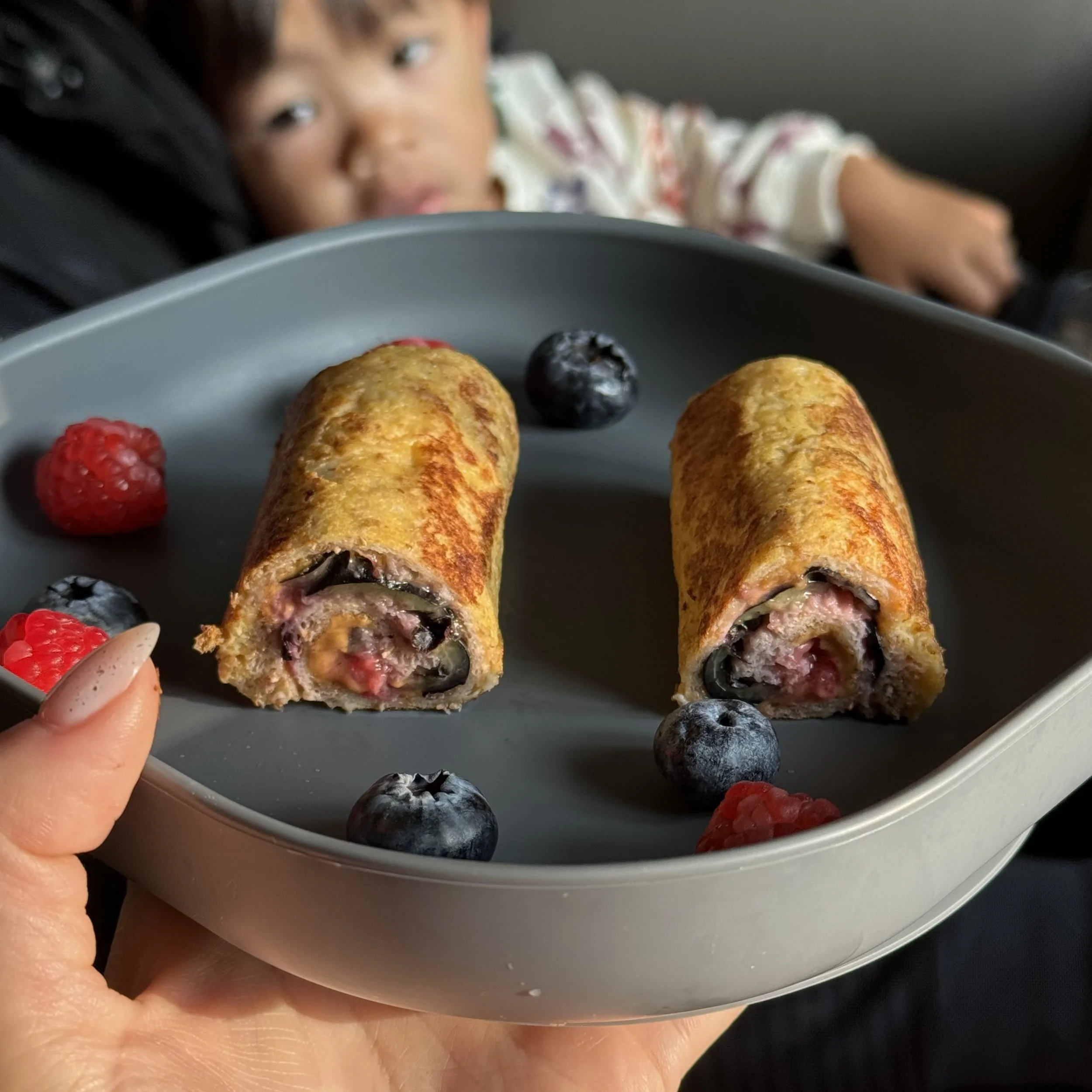 Close-up of a plate with two blueberry and raspberry-filled crepes, garnished with fresh raspberries and blueberries. In the background, a child with brown hair and a patterned shirt rests in a car seat, looking toward the camera.