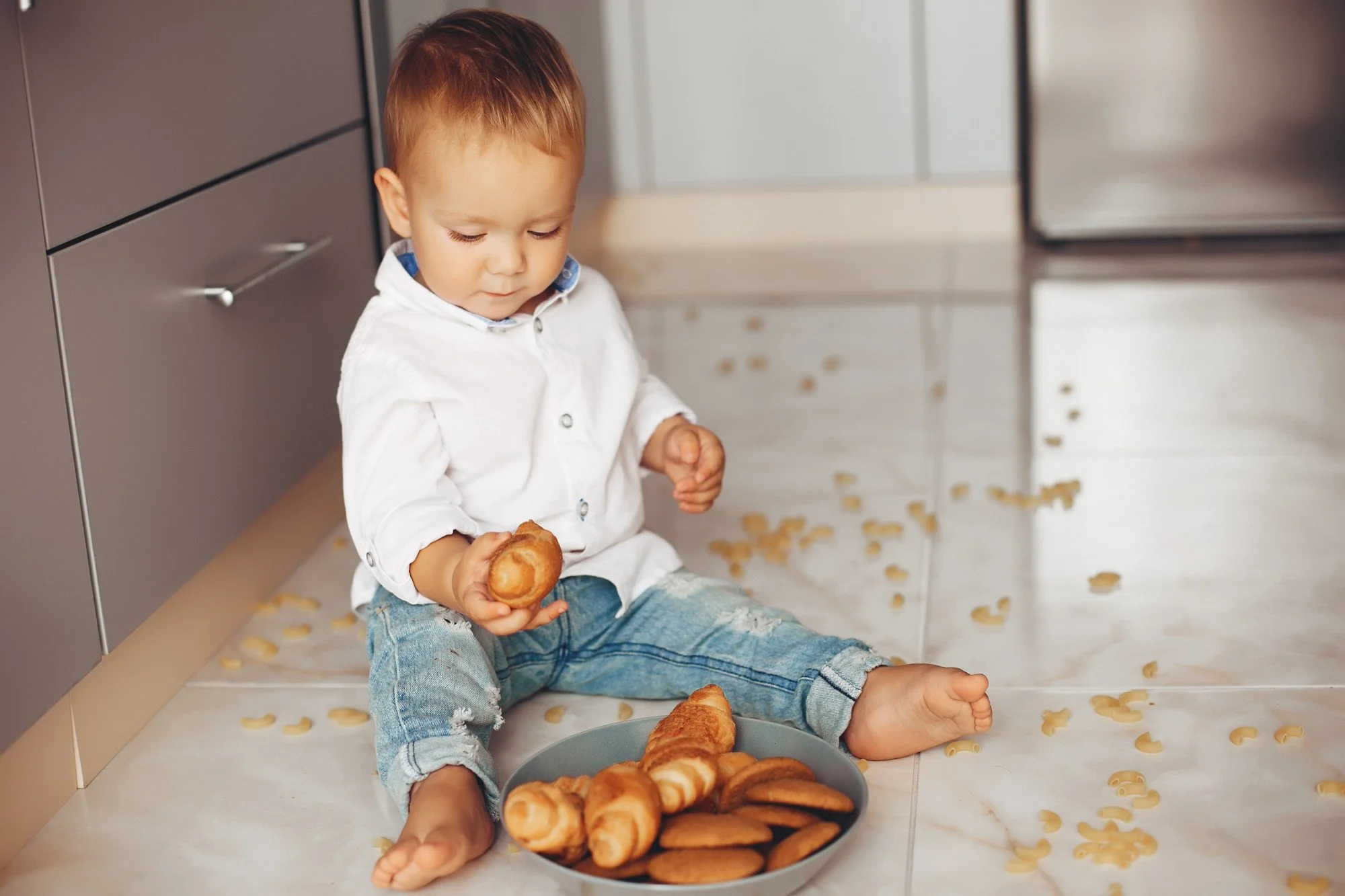 A young boy sitting on the kitchen floor, surrounded by scattered macaroni pasta, holding a bread roll, with a plate of various pastries in front of him.