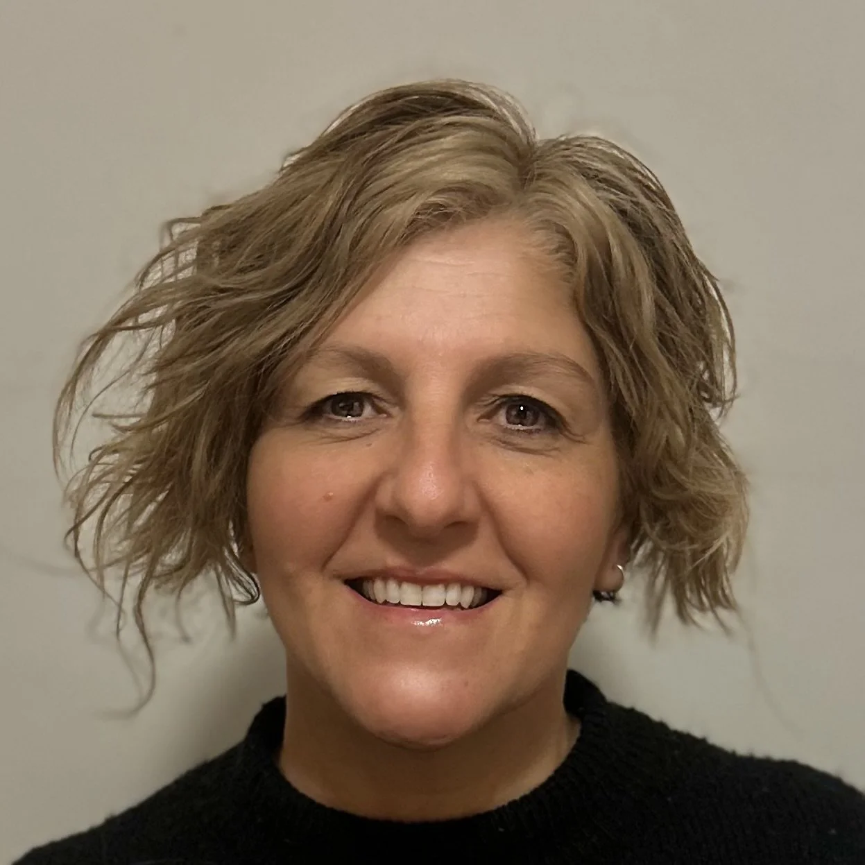 Close-up of a woman with short, wavy blonde hair smiling, wearing a black top, against a neutral background.
