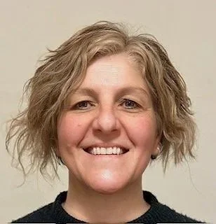 Close-up of a woman with short, wavy blonde hair smiling, wearing a black top, against a neutral background.