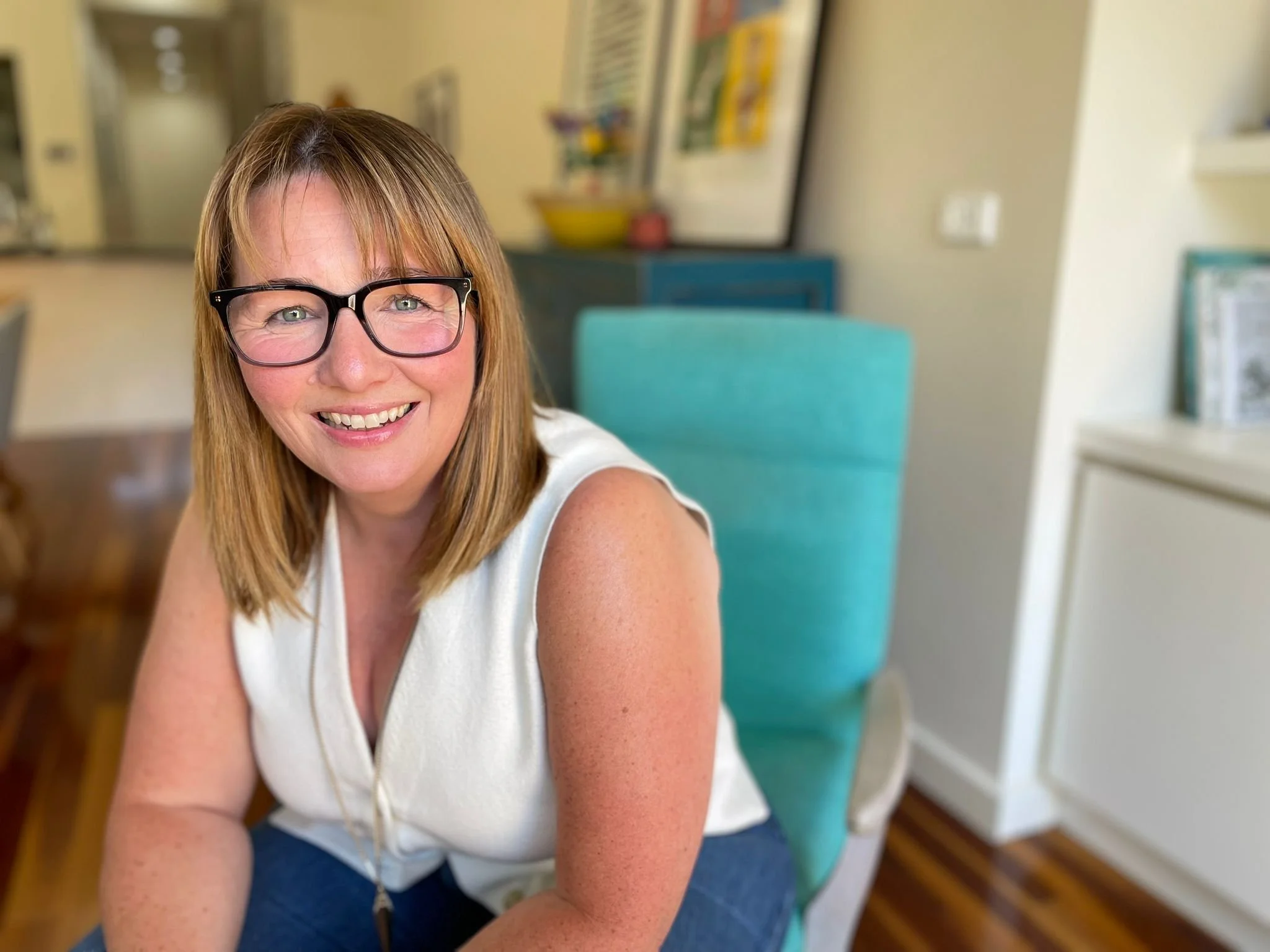 A woman with shoulder-length light brown hair, wearing black glasses and a white sleeveless top, sitting in a brightly lit room with a smile.