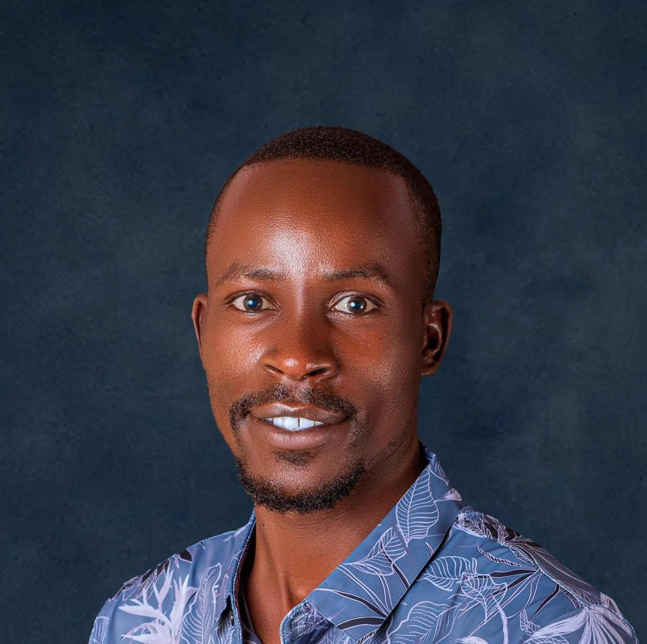A portrait of a man with short hair, a goatee, and blue eyes, wearing a patterned shirt, standing against a dark blue background.
