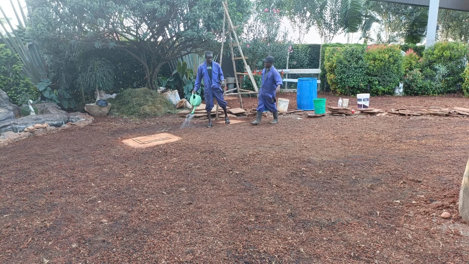 Two workers wearing blue uniforms watering a freshly laid gravel surface outdoors, with gardening tools, containers, and lush green bushes in the background.