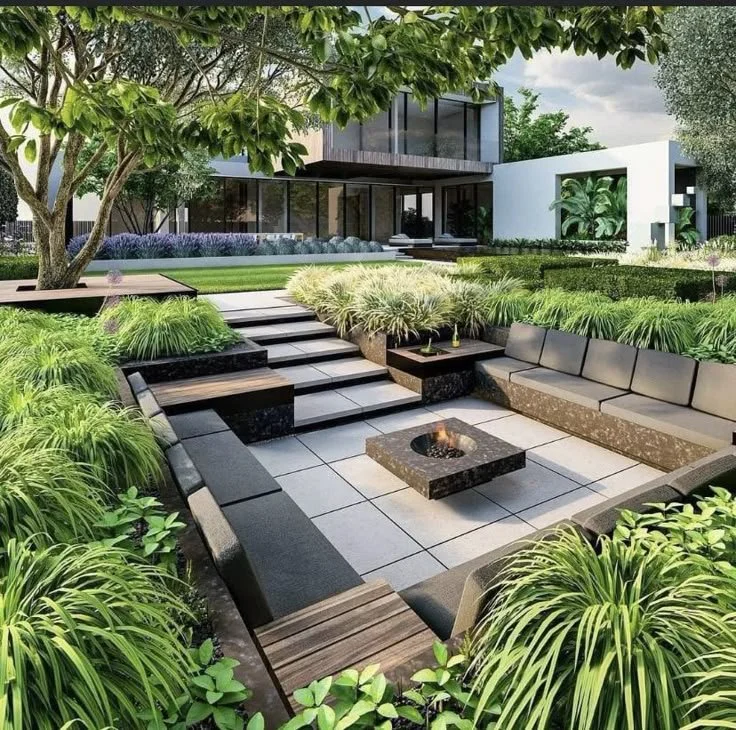 Modern backyard with steps leading to a patio, surrounded by lush green plants and trees, with a contemporary house in the background.