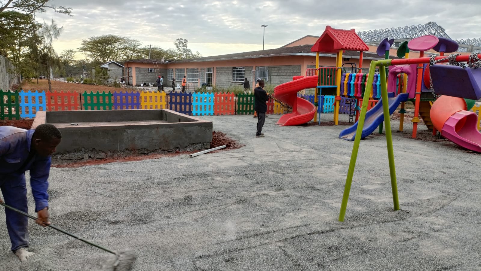 Children's playground with slides, swings, and colorful fencing, with workers and adults nearby, under a cloudy sky.
