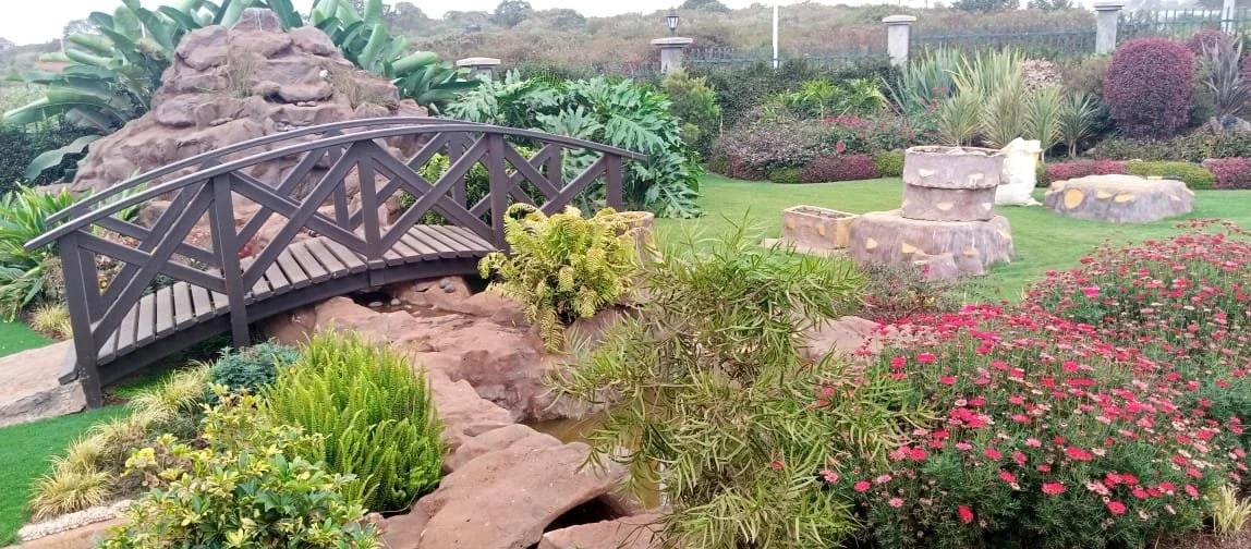 A decorative garden with a small wooden bridge over rocks, surrounded by various plants and blooming flowers, in a landscaped outdoor setting.