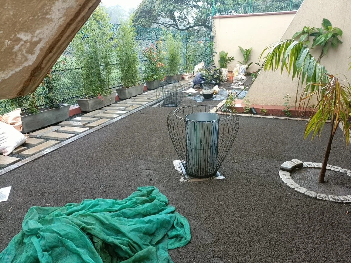 A balcony garden in construction with fresh black soil and potted plants, a green tarp on the ground, and tools and materials scattered around. A person is working near the back wall, and there are trees behind the balcony.