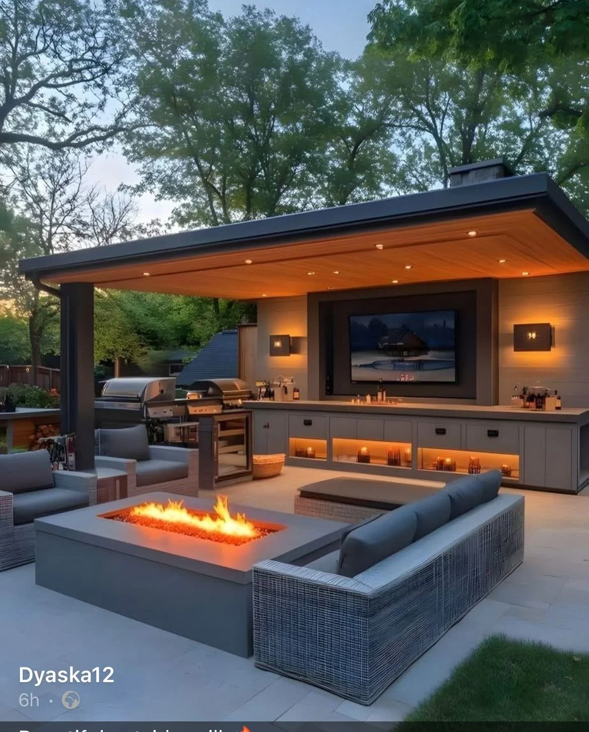 Outdoor living space with modern furniture, firepit, built-in kitchen, and a large outdoor TV, surrounded by trees.