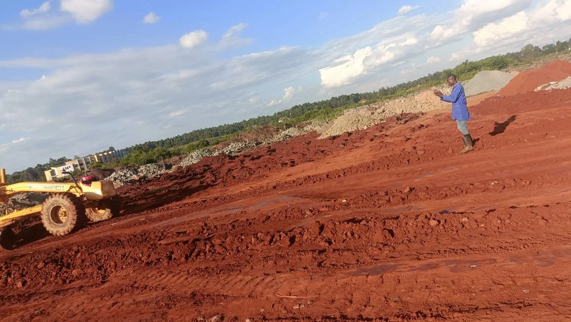 A person in a blue jacket and boots standing on a reddish dirt construction site, with a yellow bulldozer in the background and a partly cloudy sky overhead.
