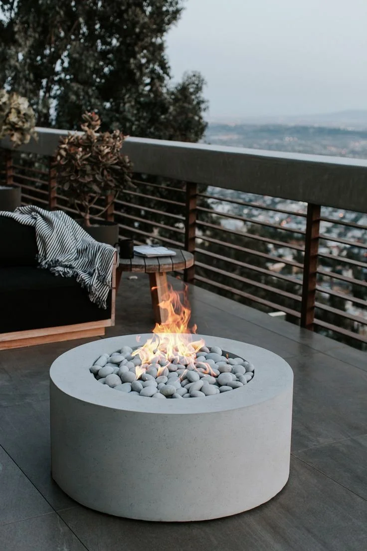 Modern outdoor patio with a fire pit filled with white stones and a small flame, a black couch with a striped blanket, a small side table with a potted plant, on a wooden deck with a metal railing overlooking a cityscape and trees in the background.