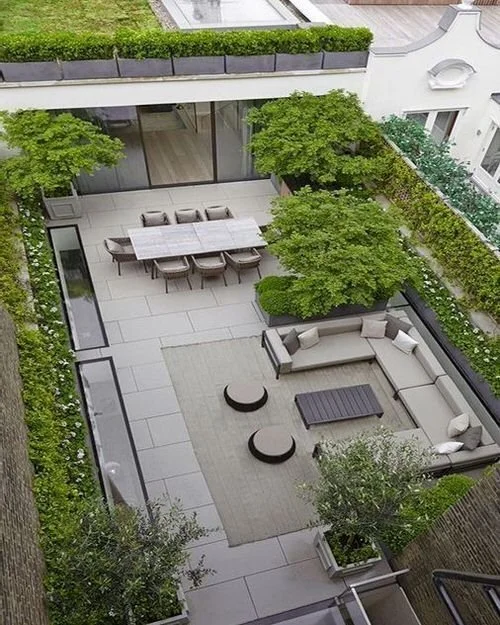 Modern outdoor patio with dining table, chairs, outdoor sectional sofa, coffee table, and greenery in planters and surrounding hedges.