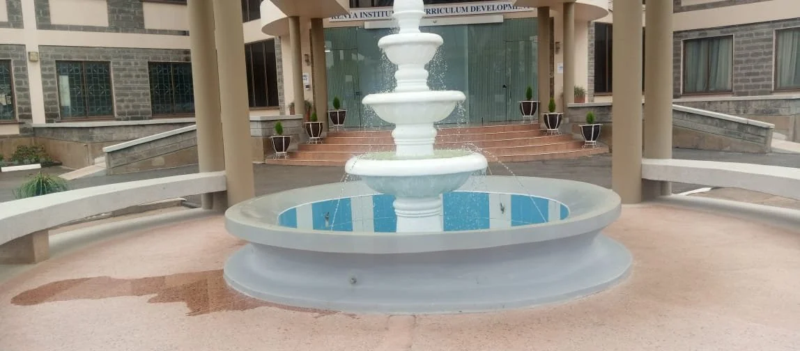 A white multi-tiered fountain in front of a modern building with steps and potted plants on each side.