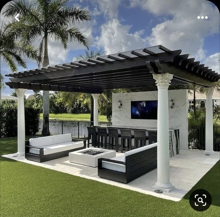 Outdoor patio with black and white seating area, a black roof with exposed rafters, white columns, a TV mounted on a white wall, a bar with multiple black chairs, and palm trees in the background.