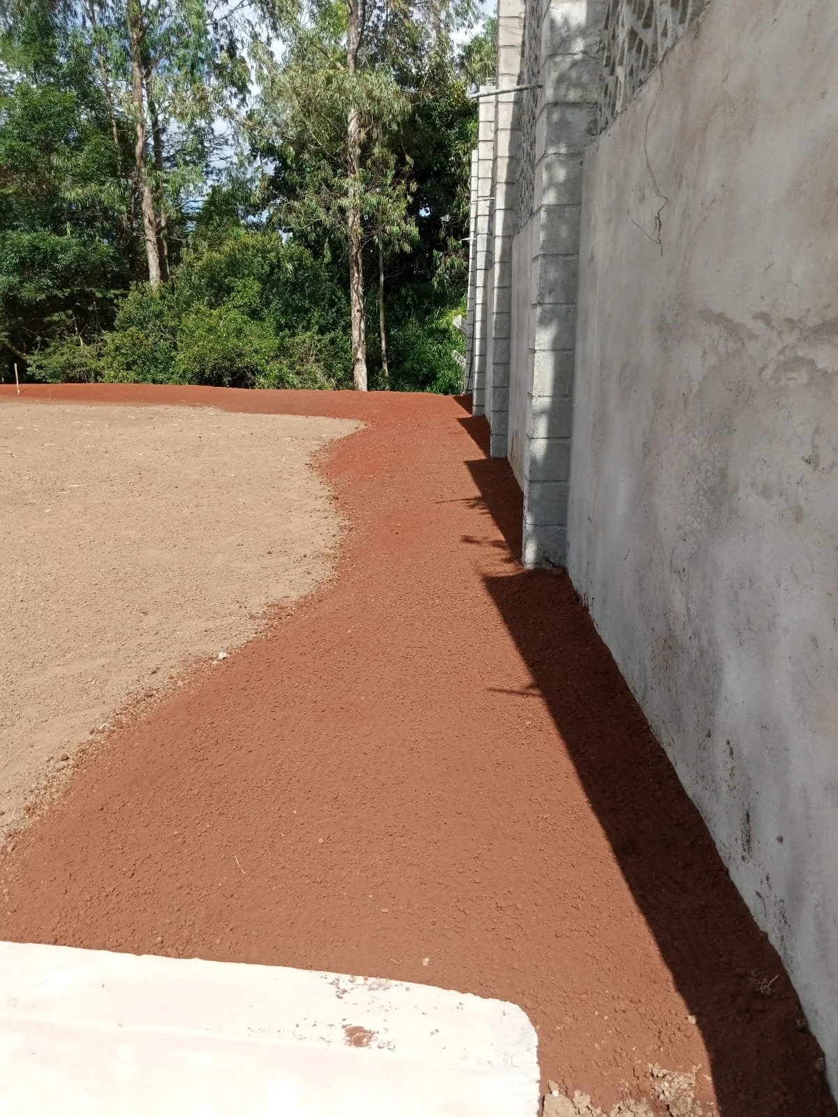 Photo shows a newly laid red gravel pathway alongside a white concrete wall, with trees visible in the background under a sunny sky.