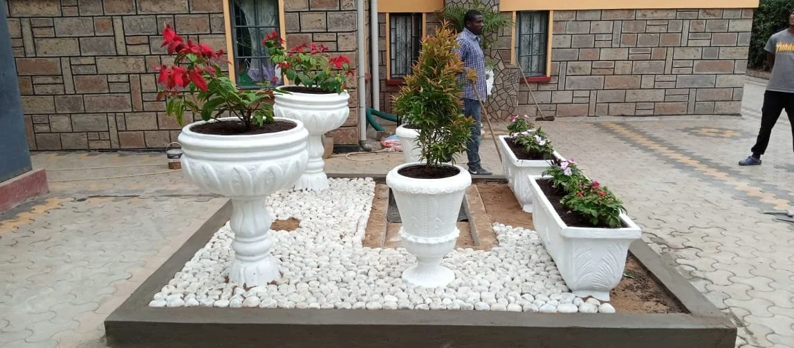 Decorative garden area with white flower pots and planters containing colorful flowers and small trees, laid with white pebbles and surrounded by paving stones.