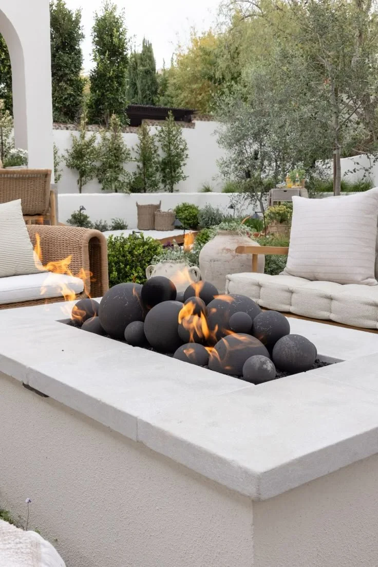 Outdoor patio with a modern fire pit filled with black stones and flames, surrounded by wicker and cushioned chairs, lush green trees, and a white wall in the background.