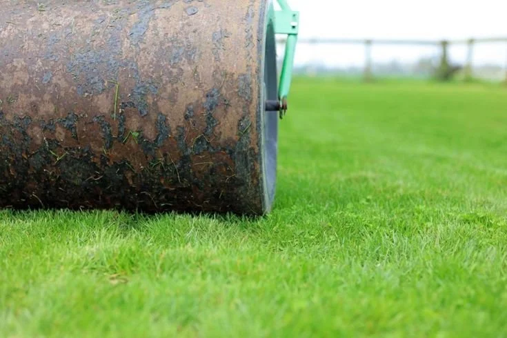 Close-up of a large rusty yellow roller on a green grass field with a white fence and cloudy sky in the background.