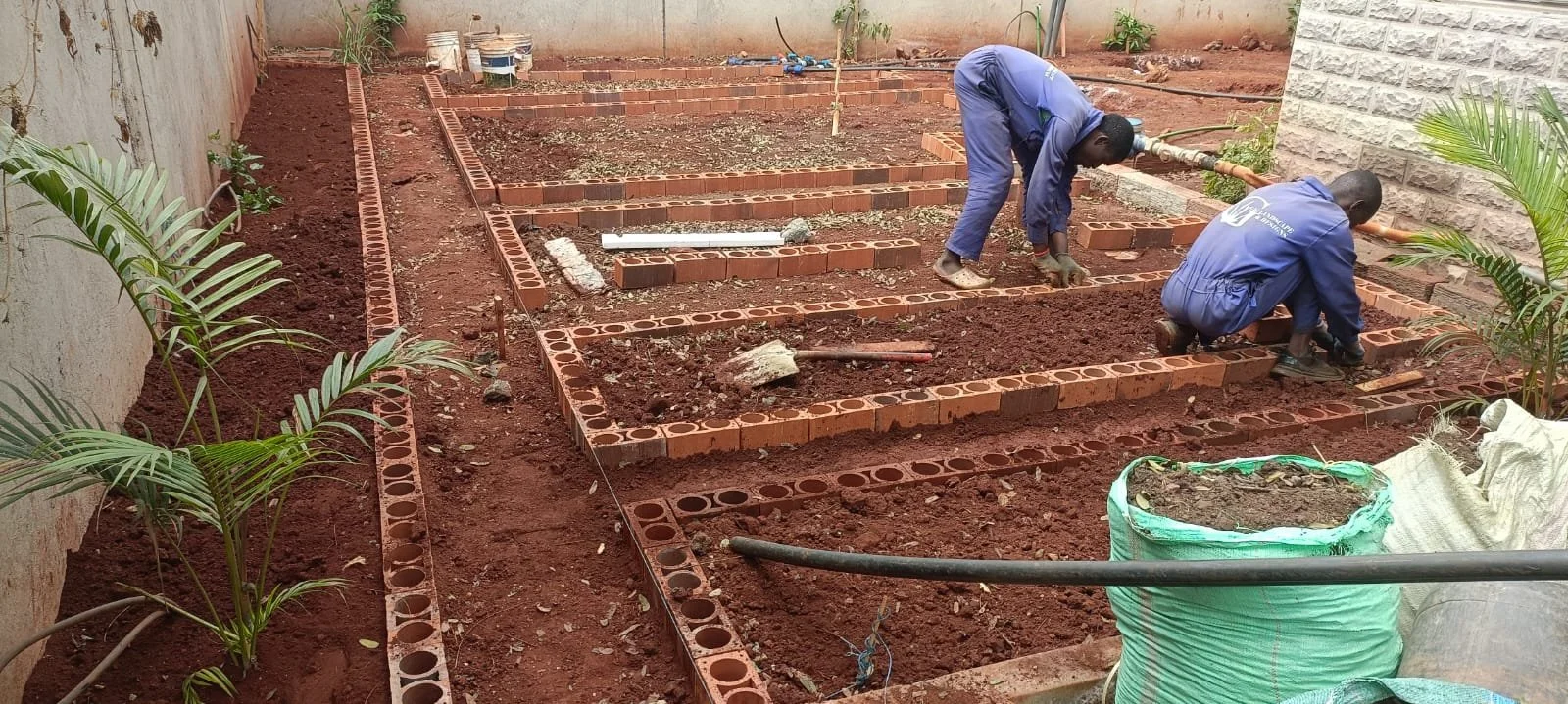 Two workers building garden bed structures with bricks, with tools and construction materials around, in a backyard or outdoor area.