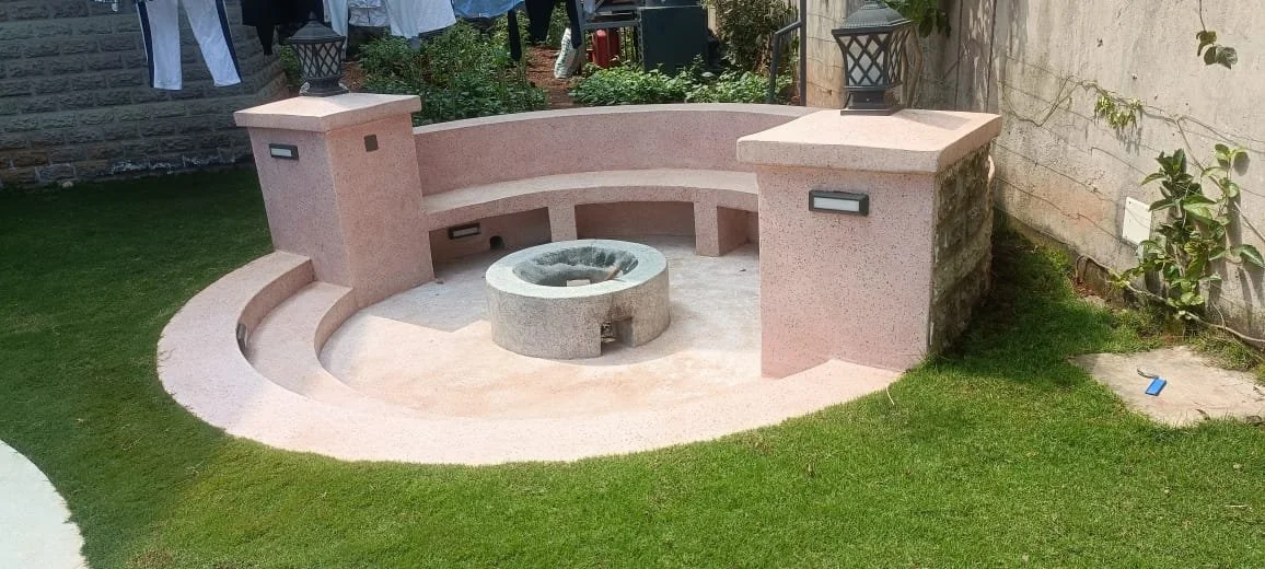 Circular outdoor seating area with a fire pit in the center, built with pinkish stone, surrounded by green grass and located beside a house wall with some plants and hanging laundry in the background.