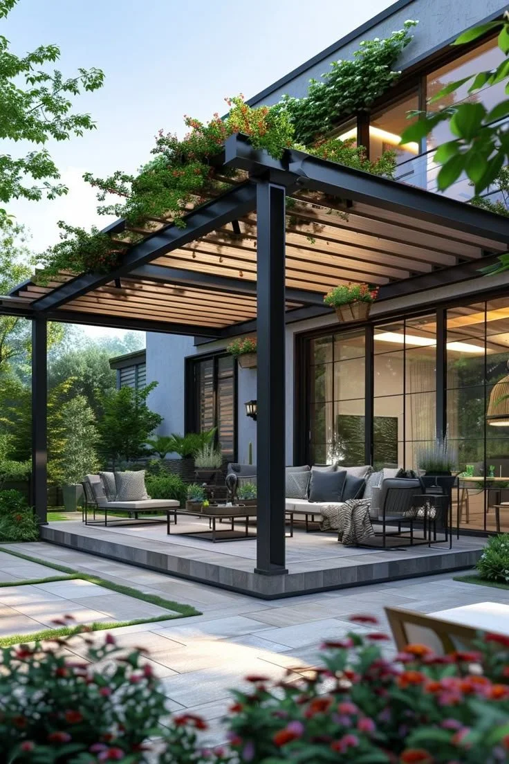 Modern backyard patio with outdoor seating and a pergola surrounded by lush greenery and flowers.