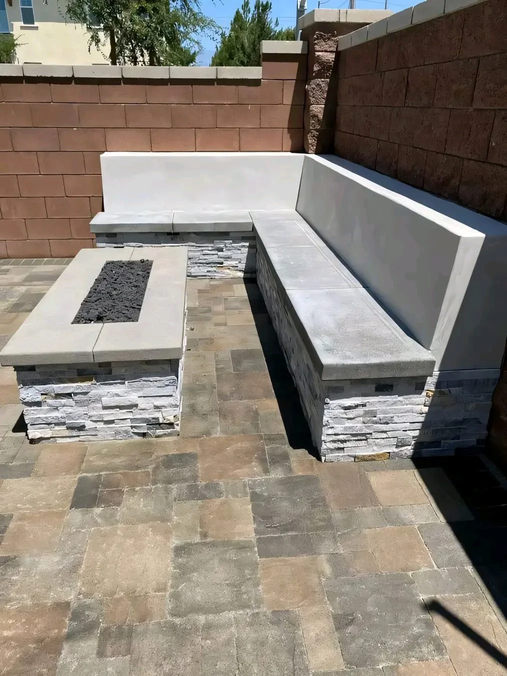 Outdoor patio seating area with stone and concrete benches arranged in an L-shape, a matching rectangular fire pit, and a brick wall in the background, on a paved stone surface.