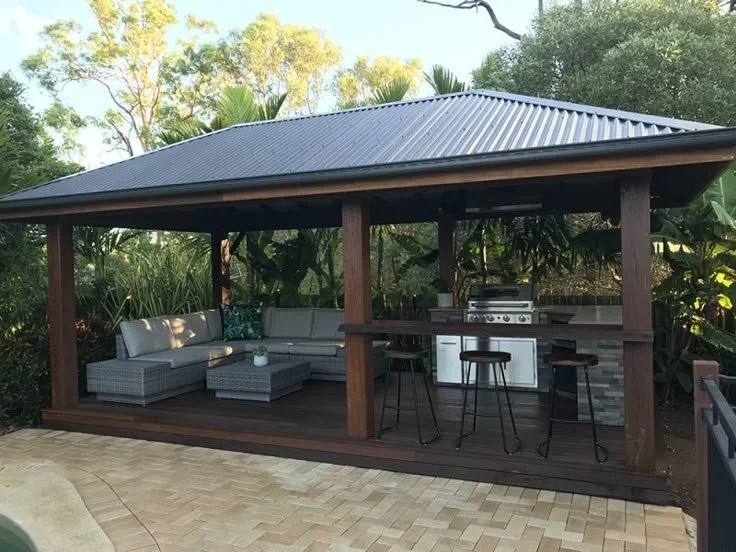 Outdoor covered patio with sitting area and built-in grill, surrounded by lush greenery.