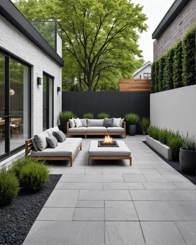 Modern backyard patio with gray tile flooring, outdoor sofa with cushions, fire pit, and potted plants, surrounded by a black fence and trees in the background.