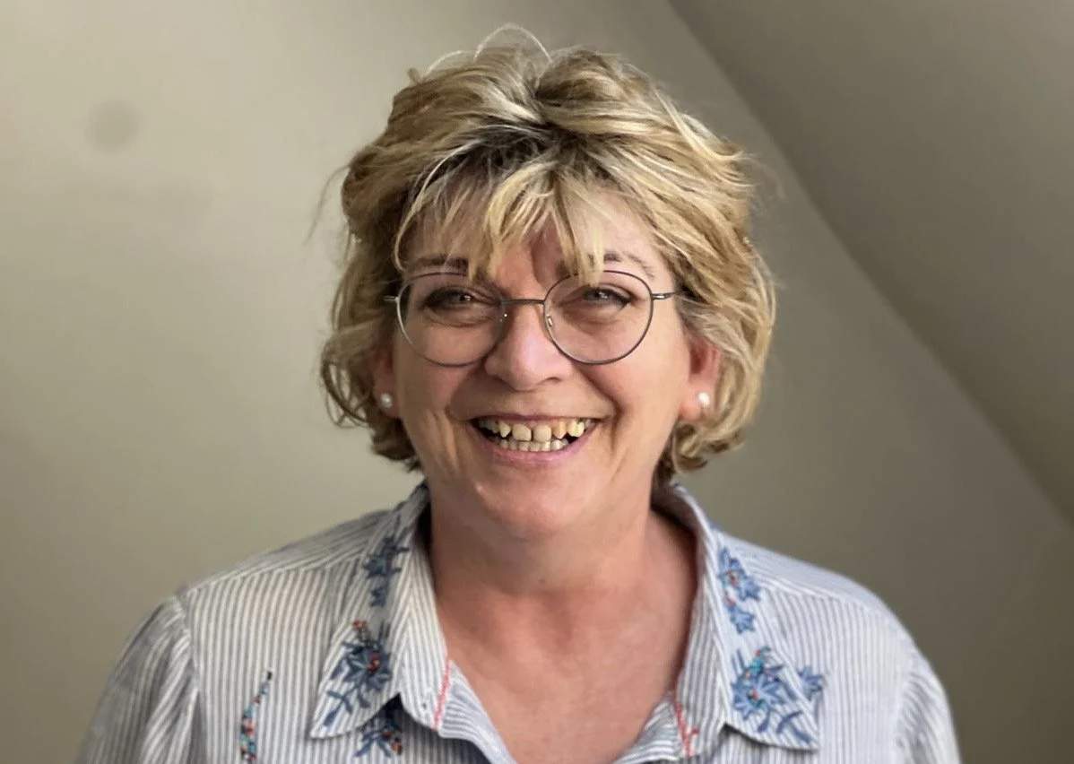 A middle-aged woman with short, curly blonde hair, glasses, and pearl earrings, smiling with visible teeth, wearing a light-colored, collared, floral-patterned shirt, standing indoors against a plain background.