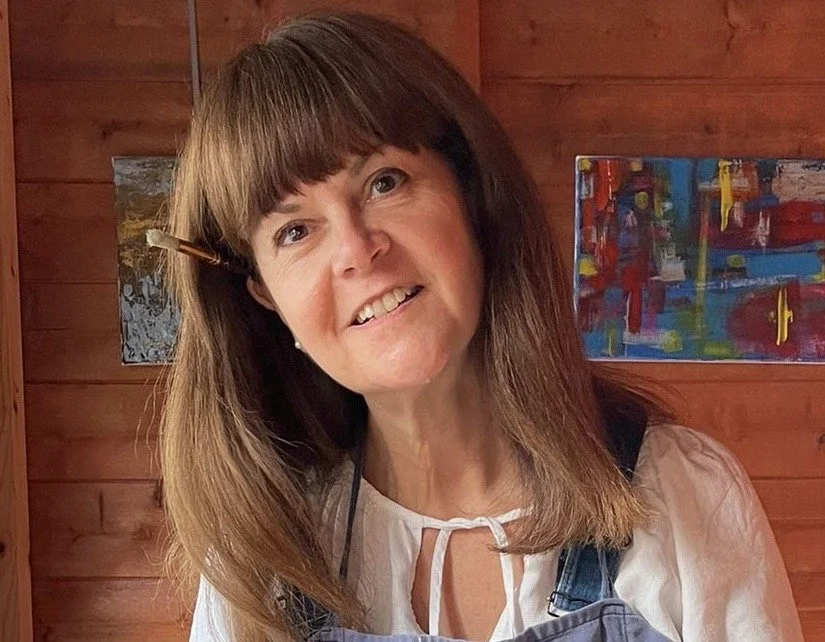 A woman with brown hair and bangs, smiling in a room with wooden walls, wearing a white top and an apron, with colorful abstract artwork in the background.