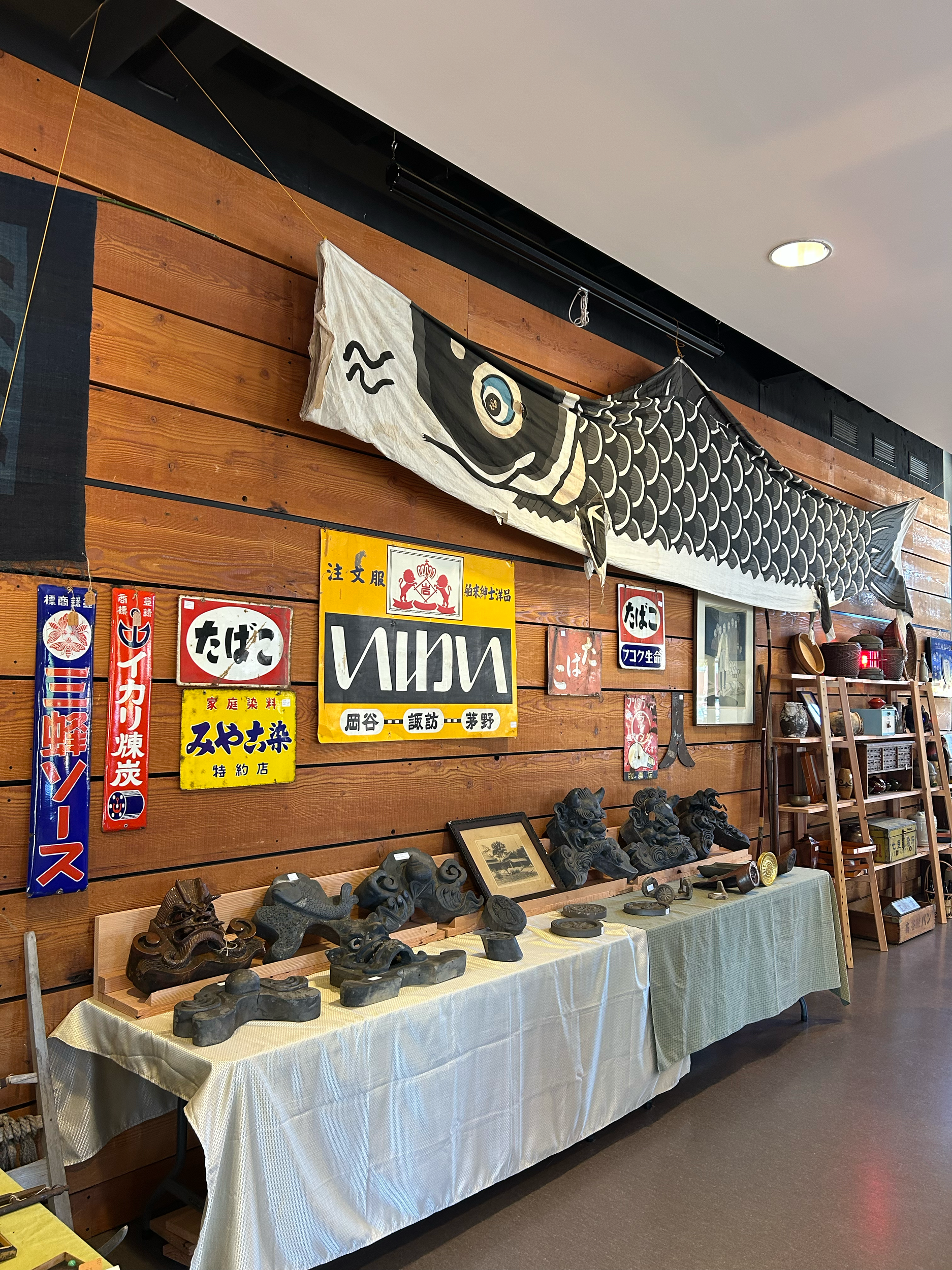Wall display featuring vintage Japanese shop signs, carved roof tiles, and a traditional koi nobori banner, reflecting commercial and architectural heritage.