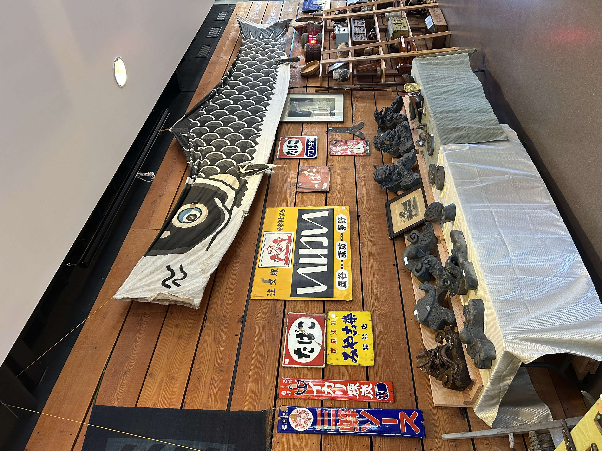 Wall display featuring vintage Japanese shop signs, carved roof tiles, and a traditional koi nobori banner, reflecting commercial and architectural heritage.