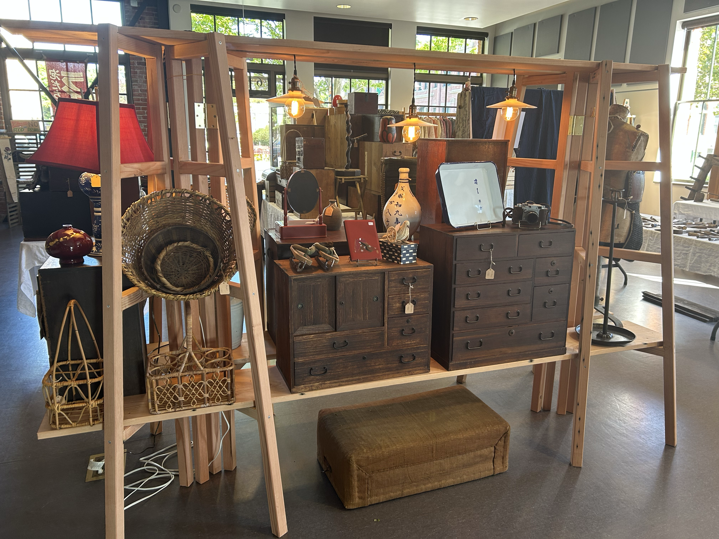 Interior display featuring Japanese tansu chests, baskets, ceramics, and period lighting, presented within a freestanding wooden shelving structure.
