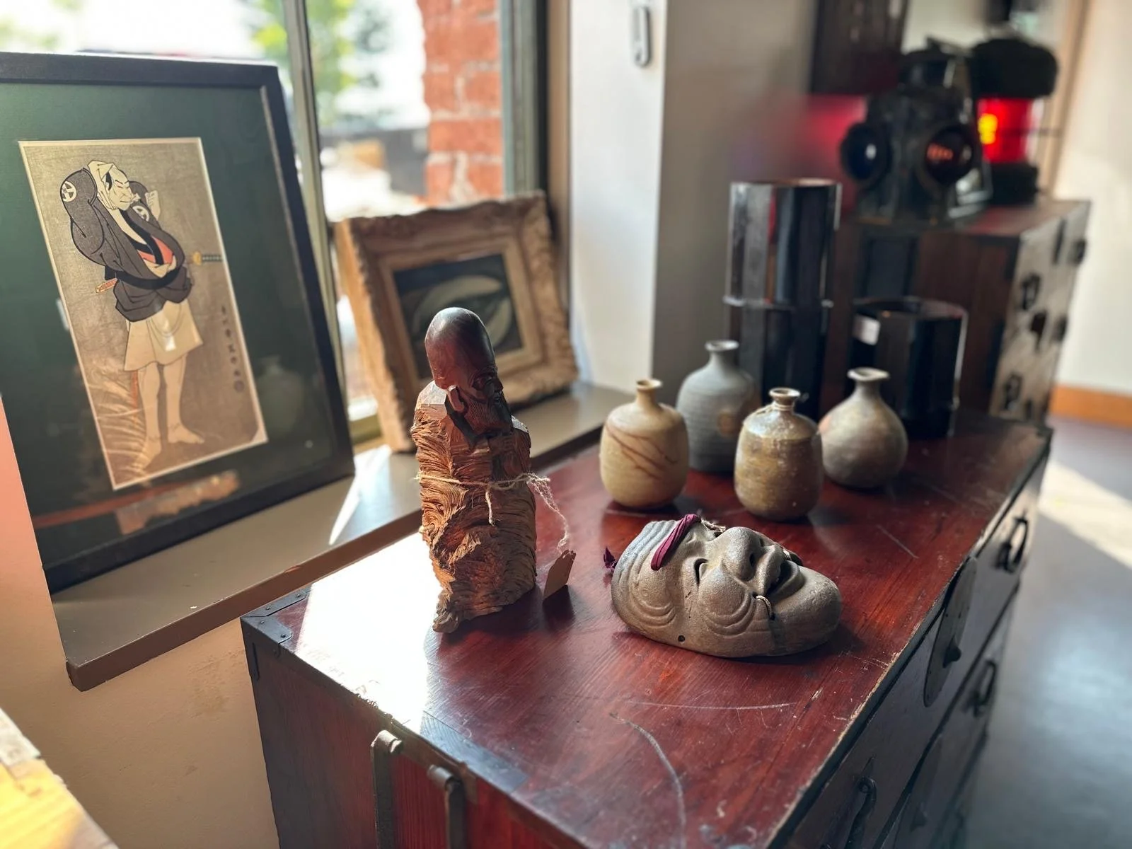 Still life of Japanese antique objects including a carved wooden figure, ceramic vessels, a noh theatrical mask, and a framed woodblock print displayed in natural window light.