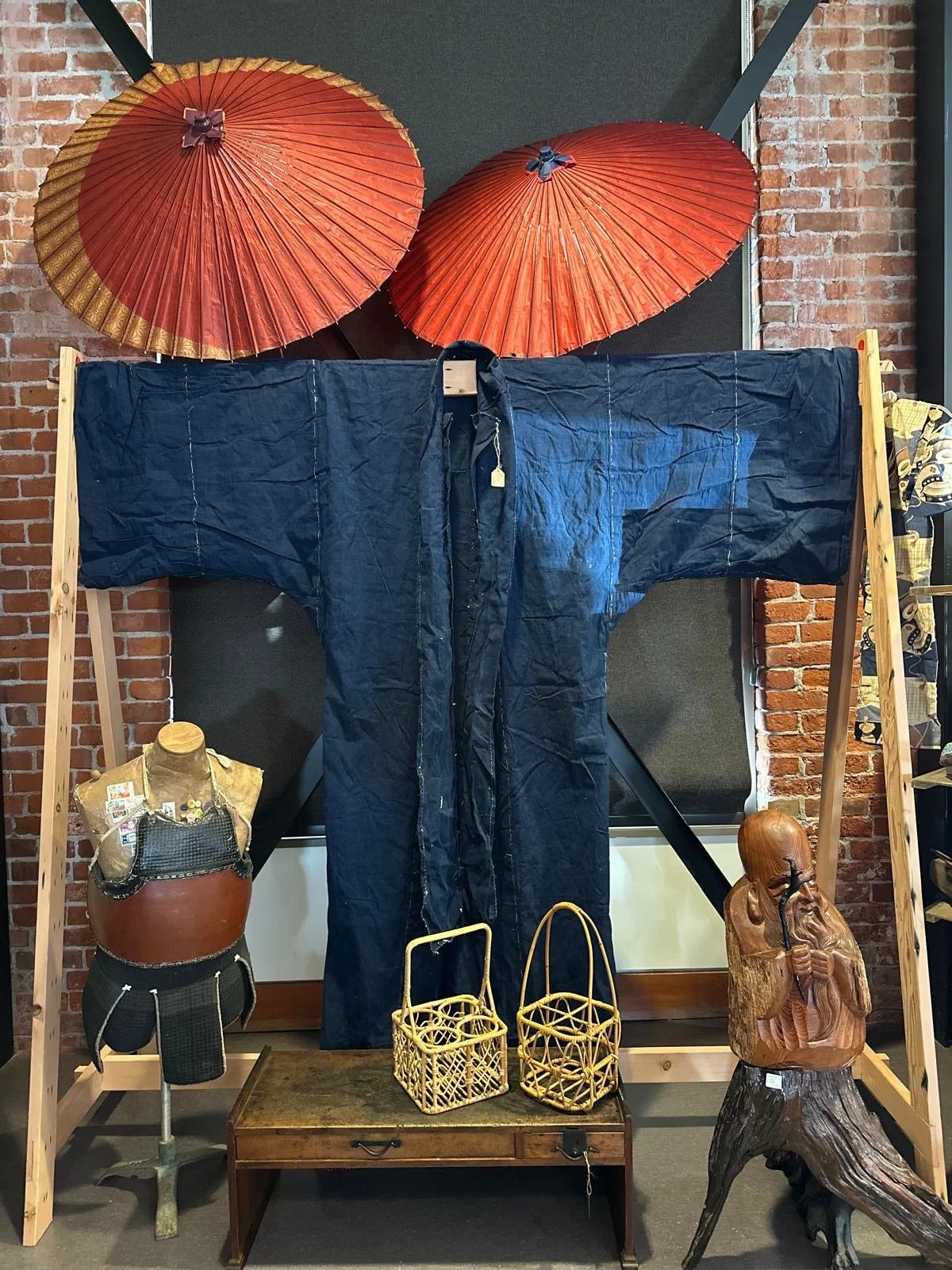 Display of a Japanese indigo-dyed garment presented with folk objects and utilitarian baskets, emphasizing traditional materials, hand stitching, and wear from use.