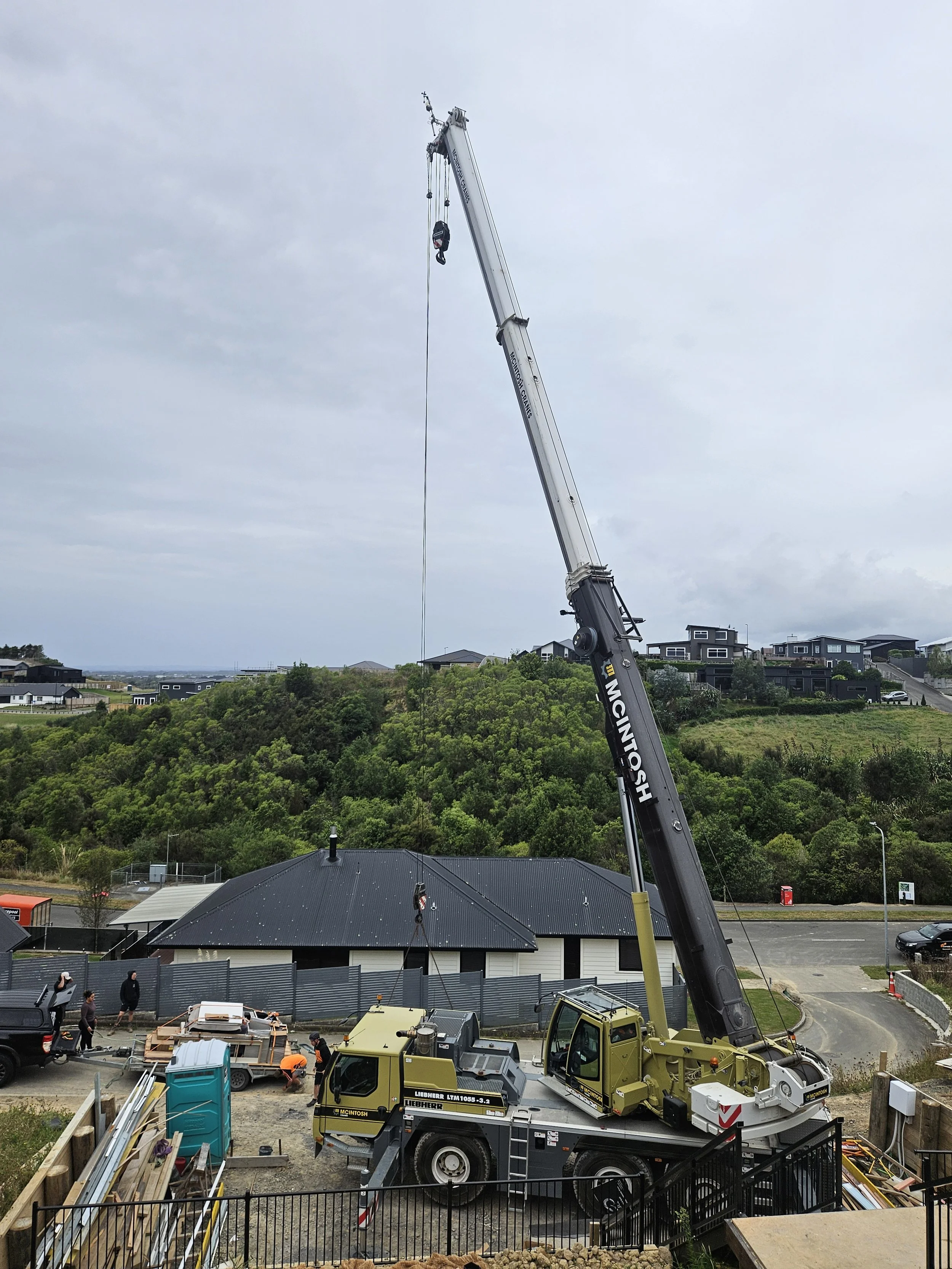 Construction site with a large yellow crane, workers, temporary fencing, and equipment, with residential houses and green trees in the background under an overcast sky.
