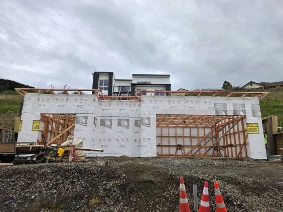 Construction site of a building with wooden framing and wrapped exterior, with orange traffic cones in the foreground and a cloudy sky overhead.