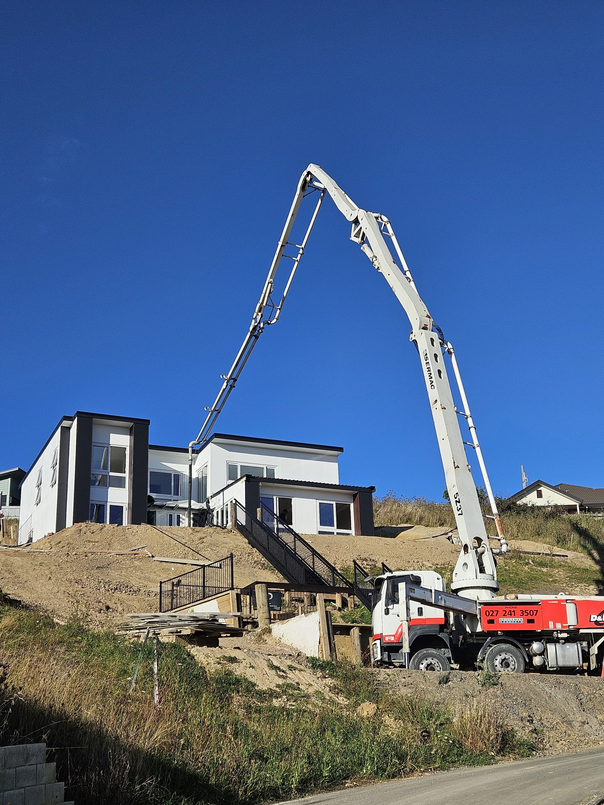 Construction site with a large crane truck lifting building materials near modern multi-story houses on a hillside under a clear blue sky.