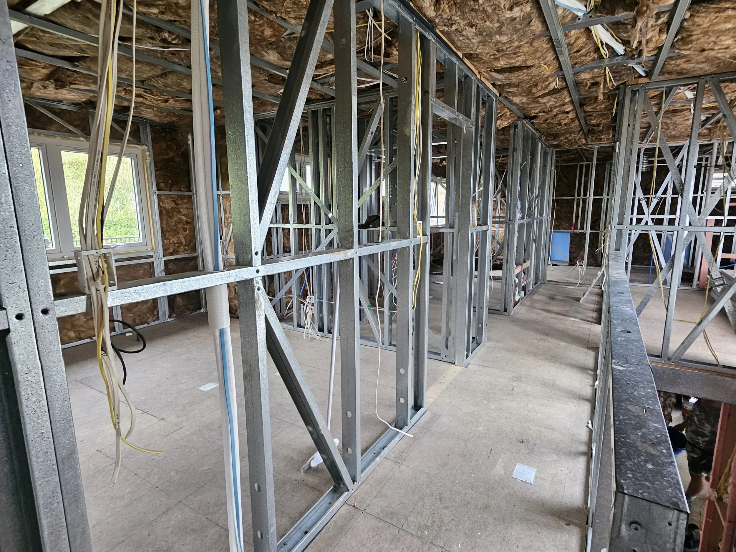 unfinished interior of a house under construction with exposed metal framing, electrical wiring, and insulation, with windows providing natural light.