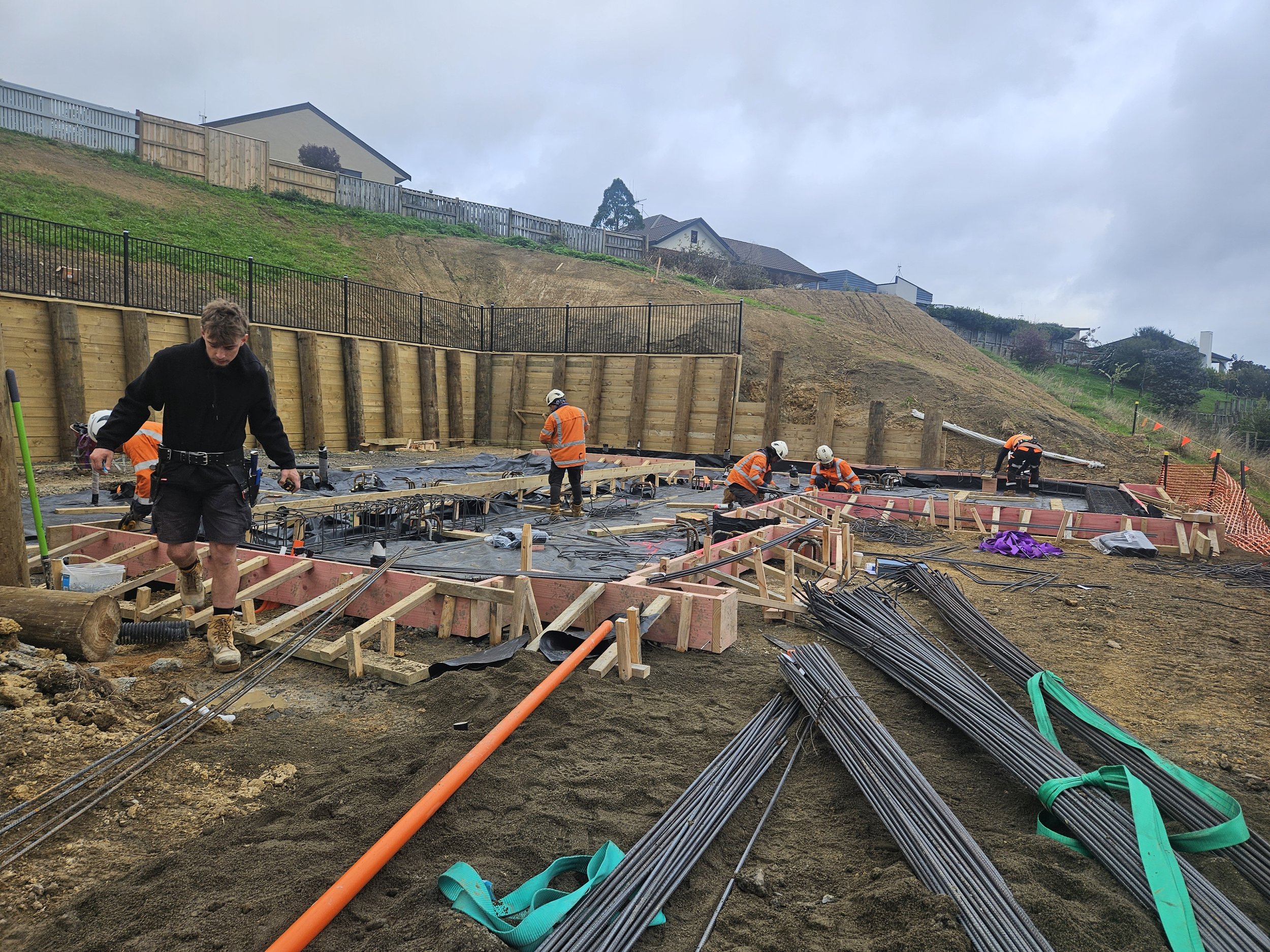 Construction workers and a supervisor working on a building foundation with rebar and wooden forms on a sloped construction site under cloudy skies.