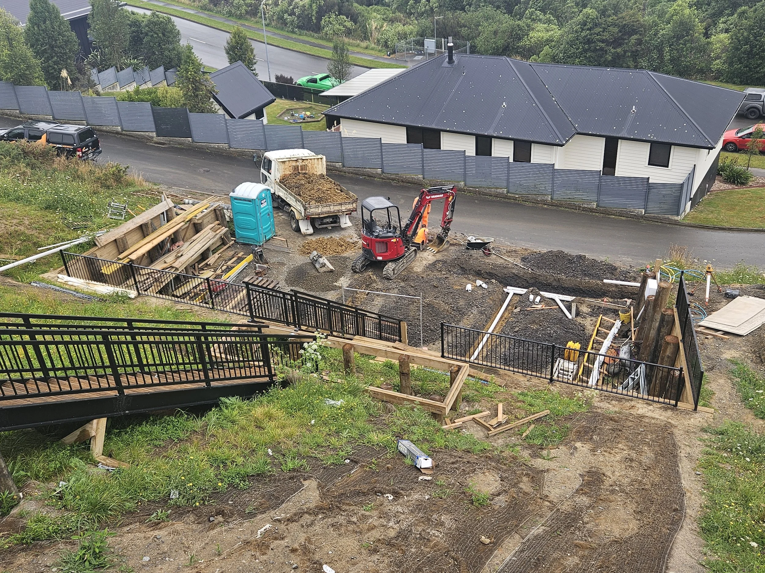 A house with a black metal roof, fenced backyard, and a construction site in the front yard. There is a small excavator and a dumped load of dirt, along with construction materials and tools. Several cars are parked nearby, and a portable toilet is on the site.