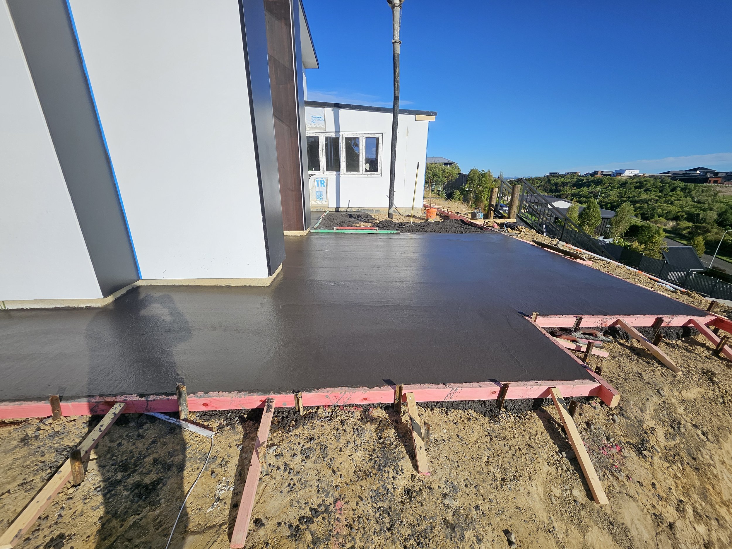 Newly poured concrete on a construction site with wooden formwork and building materials, with a background of trees and houses, under a clear blue sky.