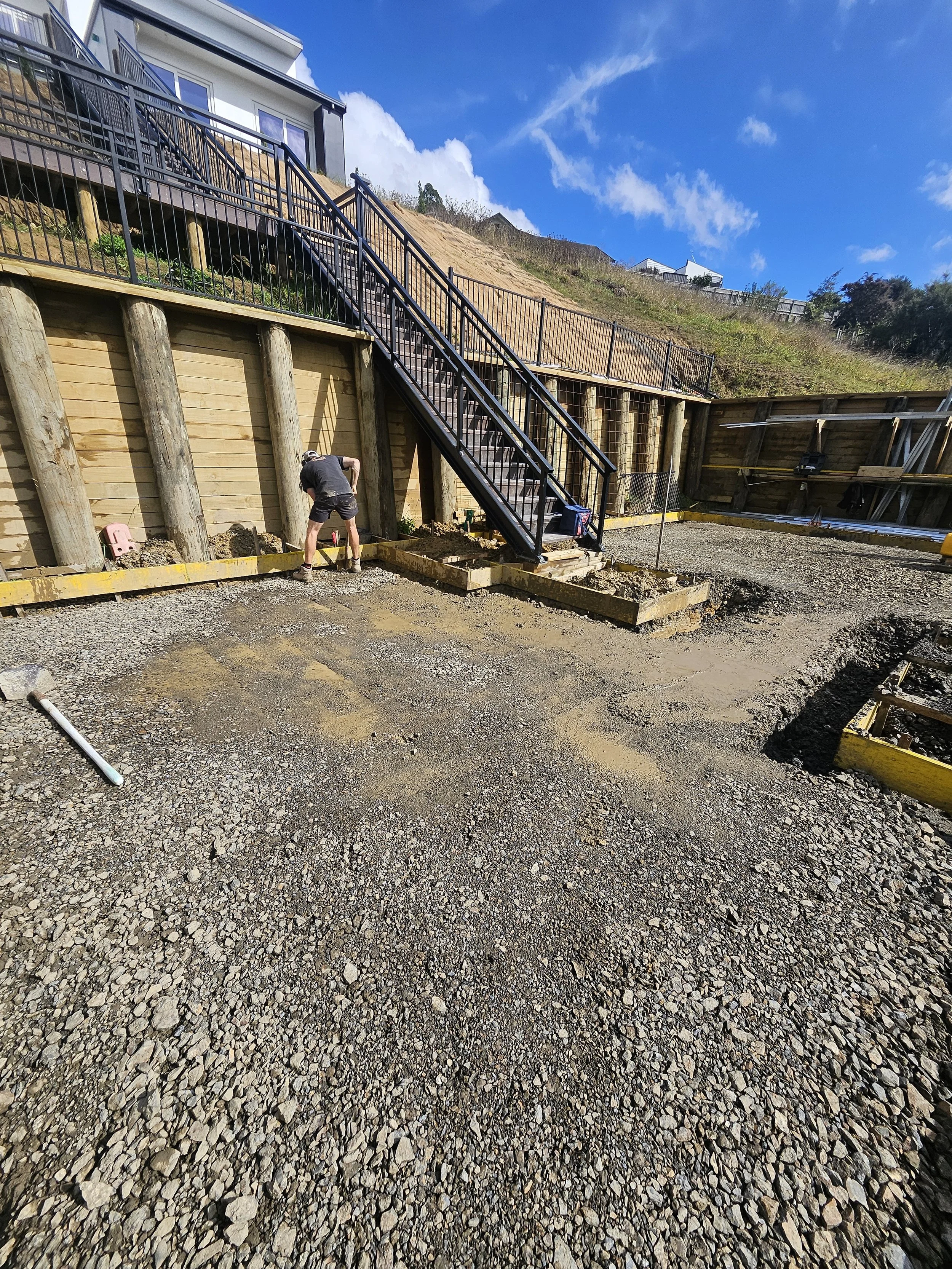 Construction site with gravel ground, a worker installing a new paved area, wooden retaining walls, and a staircase leading up to a house on a hillside under a blue sky with clouds.