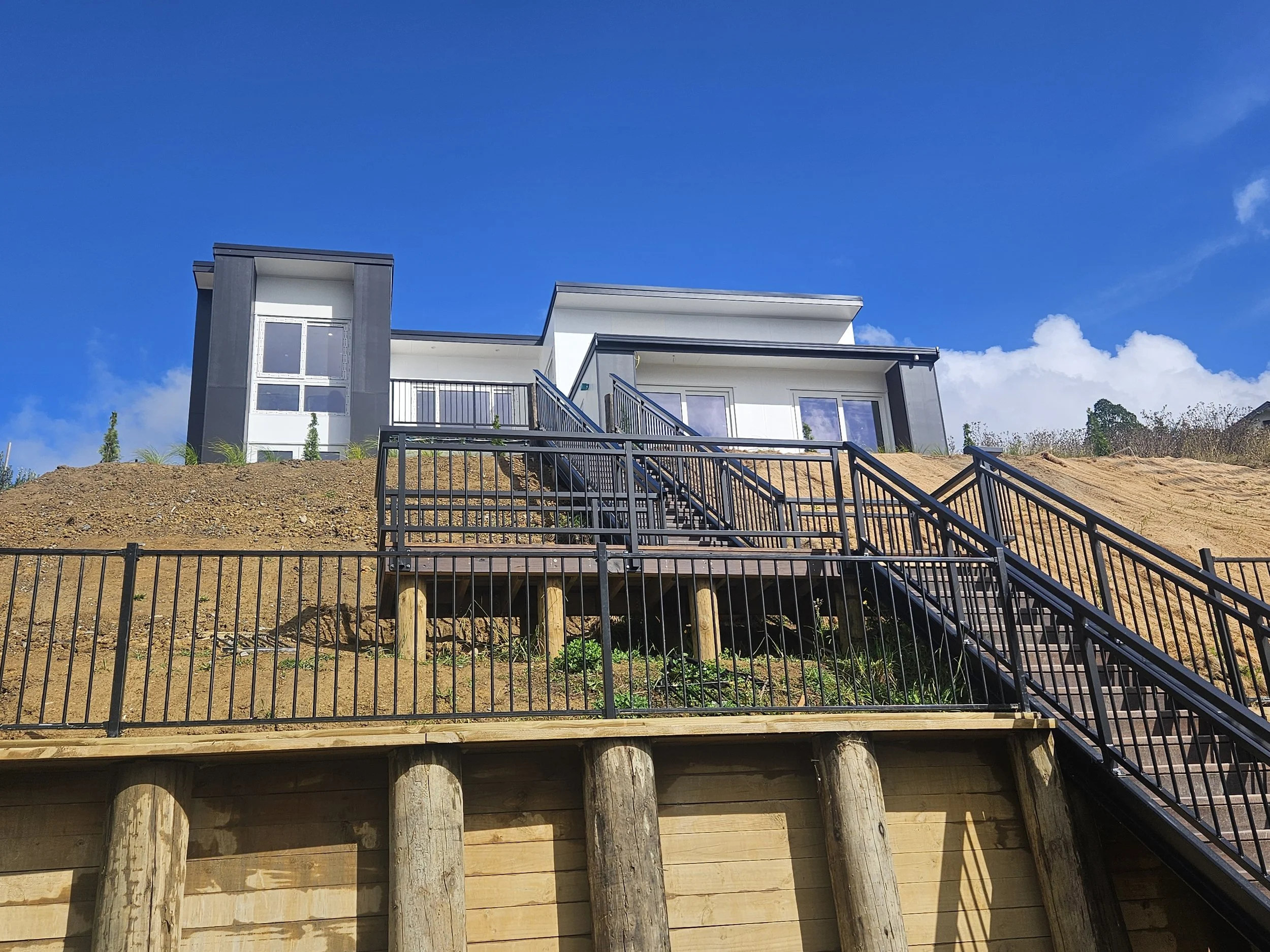 Modern two-story house with white and gray exterior, metal stairs leading to an upper balcony, and a wooden fenced basement area under construction, set against a bright blue sky with a few clouds.